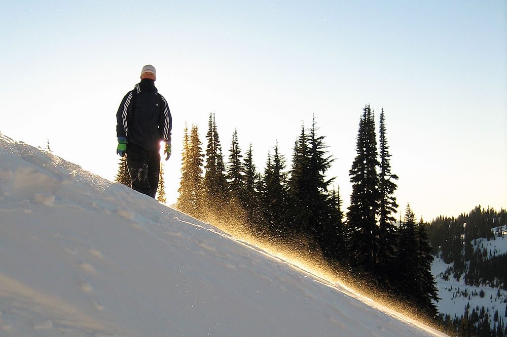 A person hikes up a snowy mountain with a backdrop of evergreen trees and a clear blue sky.