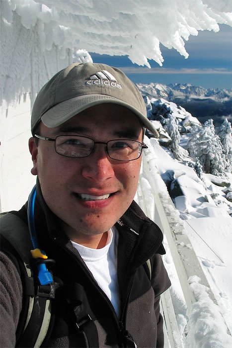 A smiling person stands on a snow-covered mountain peak, wearing glasses and a backpack.