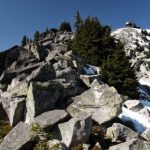 Cheer Bear A rocky mountain summit with patches of snow, surrounded by evergreen trees under a clear blue sky.