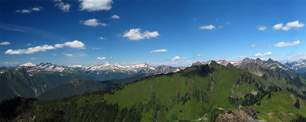 A breathtaking panorama showcasing snow-capped mountain peaks against a backdrop of lush green hills under a vibrant blue sky with scattered clouds.