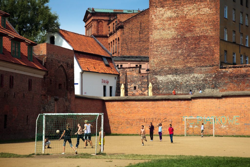 A group playing soccer on a dirt field surrounded by old brick walls.