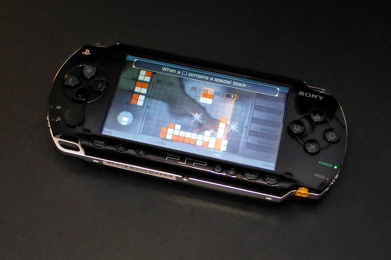 A close-up of a Sony PSP gaming device displaying a Tetris game. The portable console is set against a dark background, highlighting its sleek design and vibrant screen.