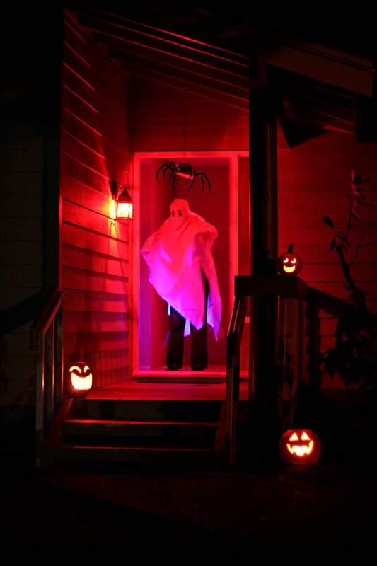 A haunted porch lit in red features a glowing ghost in the doorway and carved jack-o'-lanterns on the steps, creating a festive and eerie Halloween scene.