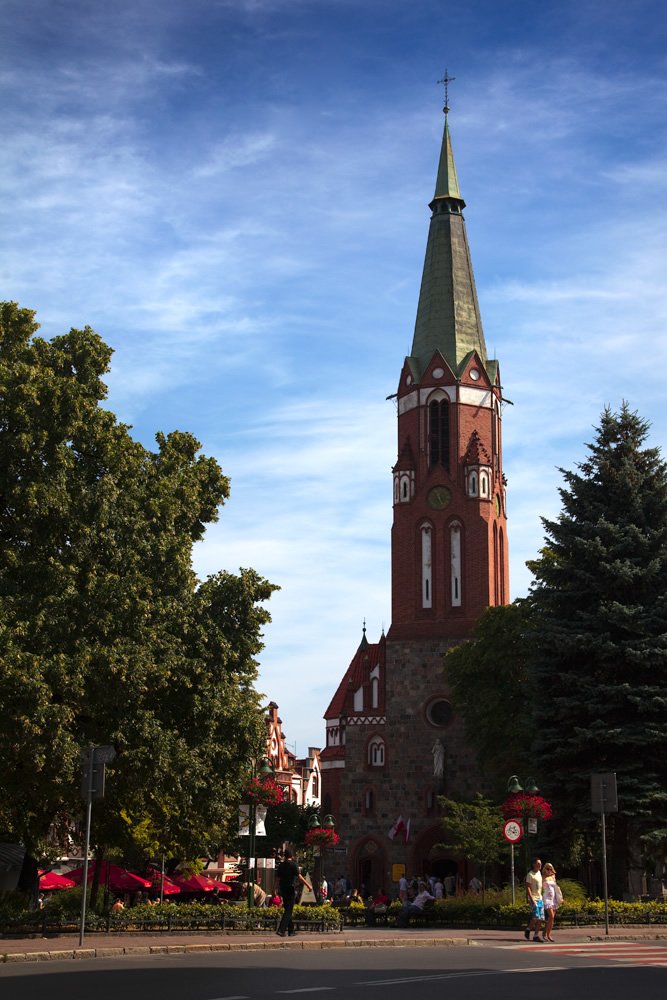 A tall church with a pointed spire stands against a clear sky in Vilnius, Lithuania. The scene includes surrounding trees and people walking nearby.