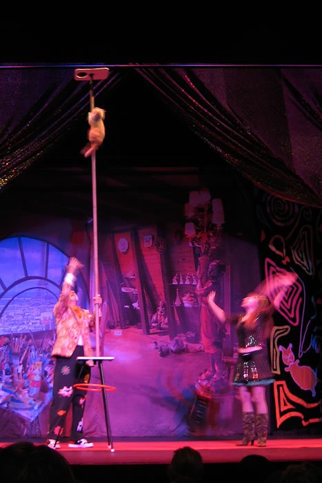 A vibrant stage setup with performers and dramatic lighting, featuring a pole and colorful props.
