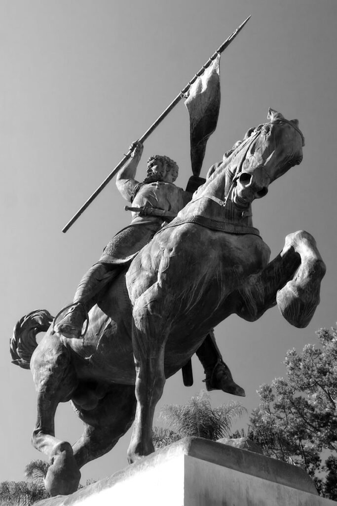 A striking black and white image showcases a dramatic statue of a medieval warrior mounted on a rearing horse. The warrior holds a tall flagpole aloft, symbolizing valor and strength.