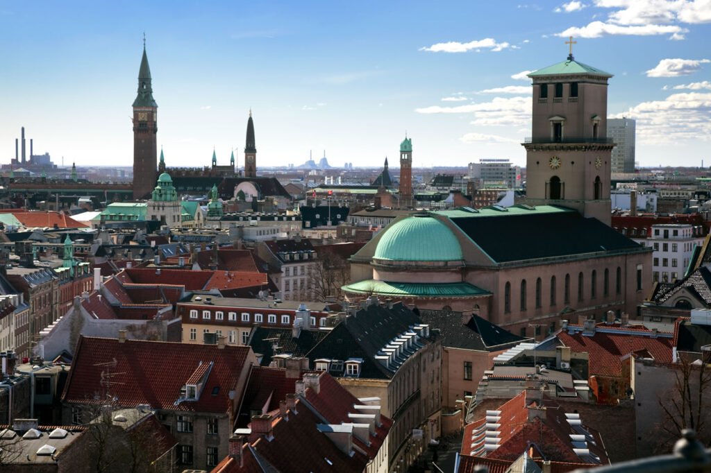 panoramic view of Stockholm cityscape featuring red-tiled roofs, green domes, and historic church spires under blue sky