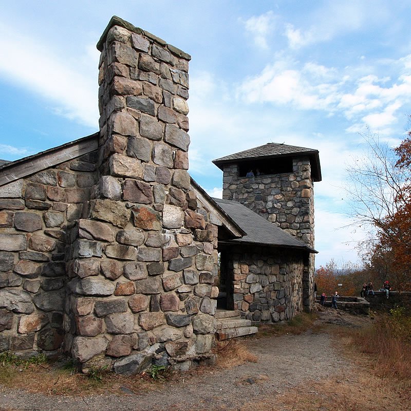 A stone cabin surrounded by autumn trees under a cloudy sky. A gravel path leads to the entrance of the rustic structure.