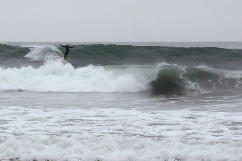 A surfer wears a wetsuit while riding a wave on an overcast day. The ocean is rough with gray clouds above.