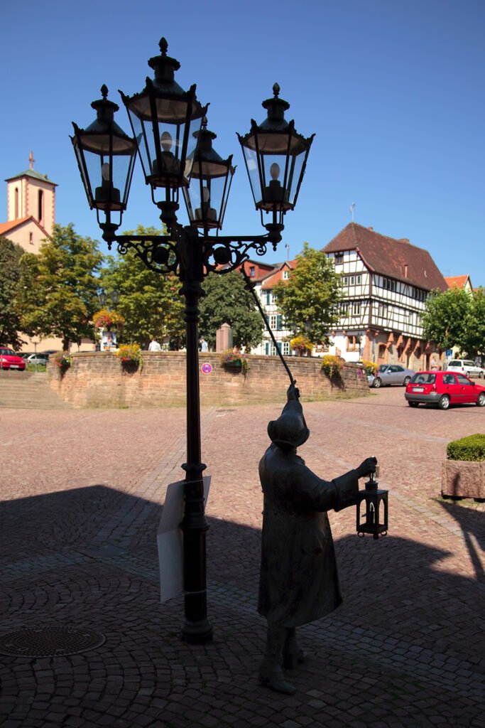 A statue of a person lighting a historic lamp against a blue sky. Traditional buildings and a red car are in the background.
