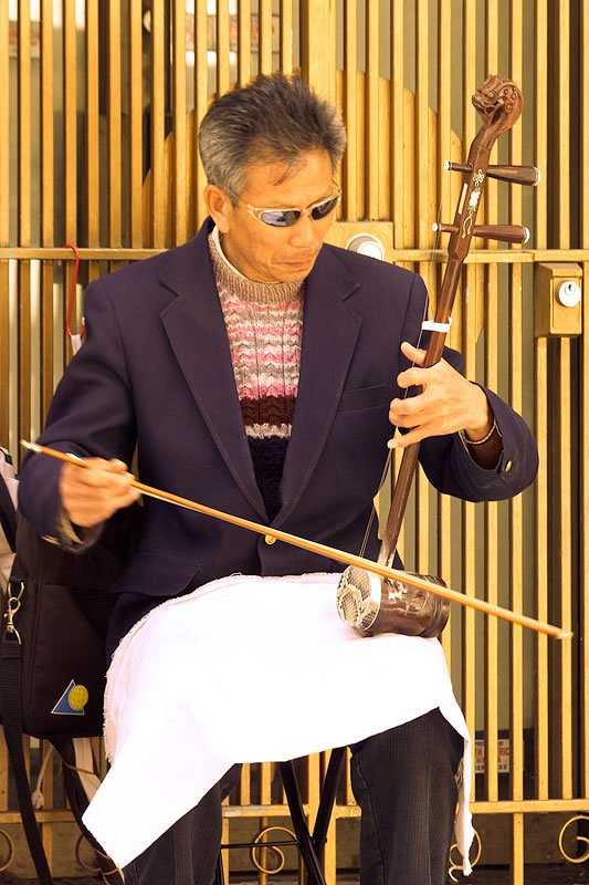 A man in a blue suit plays a stringed instrument on the street. He is focused and seated against a golden slatted background.