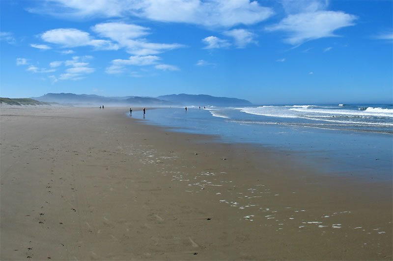 A panoramic view of a beach on a clear day, featuring sandy shores and ocean waves under a blue sky with scattered clouds.