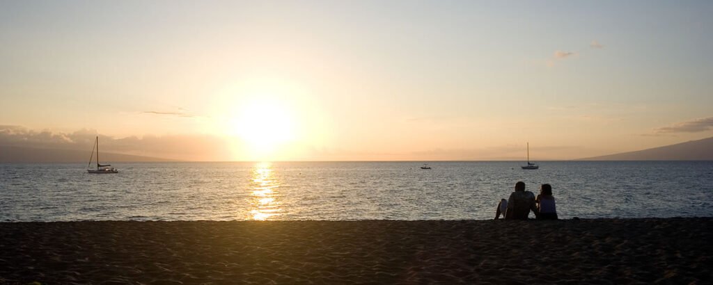 A couple enjoys a quiet moment on the beach as the sun sets over the water, casting a warm golden glow across the scene.