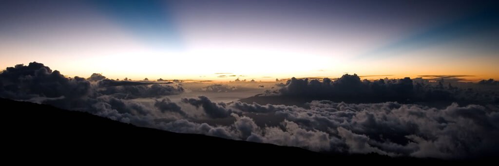A stunning sunset scene with golden hues above a sea of clouds. The horizon is lined with dark clouds, creating a dramatic contrast.