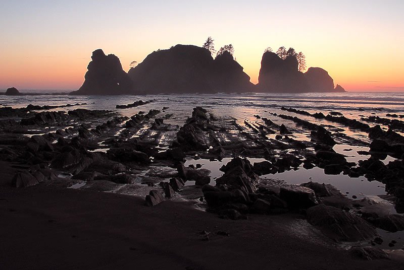 A serene sunset over the iconic sea stacks at Cannon Beach, Oregon, casting a warm glow across the Pacific Ocean and rocky shoreline.