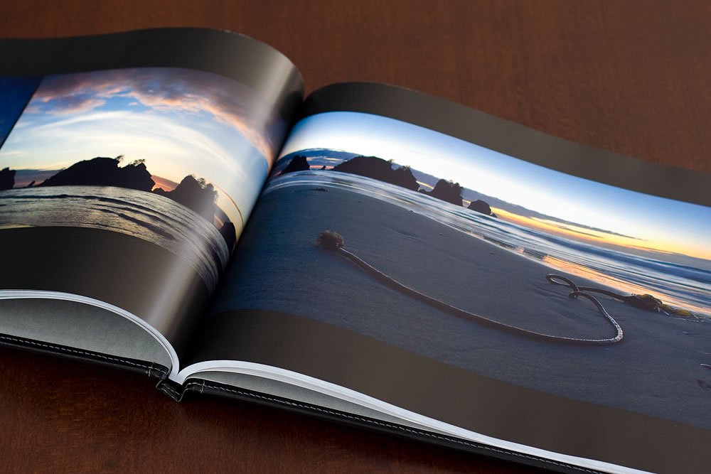 A photo album open to a double-page spread showing a sunset over the ocean and a heart drawn in the sand. The setting reflects Pacifc Northwest vibes of John & Kristie.