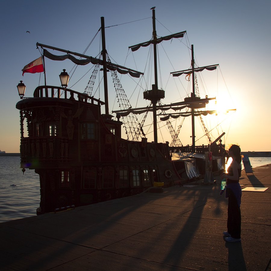 A ship's silhouette against a golden sunset, with a person standing nearby on the dock.