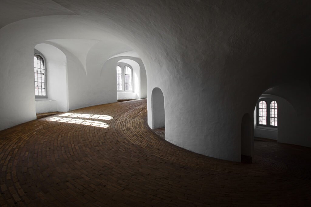 A surrealistic view of a curved interior space with arched windows and sunlight streaming in, creating patterns on the textured floor.