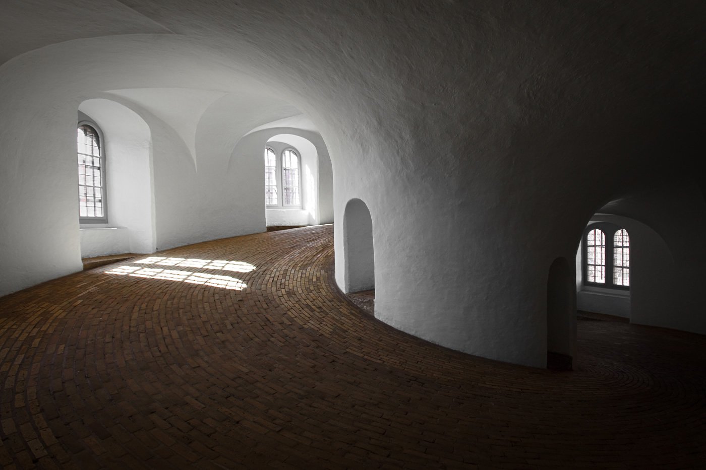 Rundetaarn A surrealistic view of a curved interior space with arched windows and sunlight streaming in, creating patterns on the textured floor.