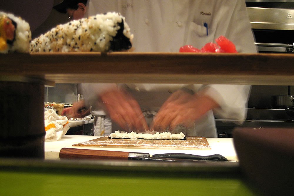 A sushi chef skillfully prepares rolls in a kitchen setting, with various ingredients visible on the counter.