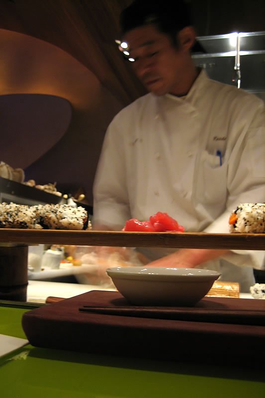 A chef in a white uniform is preparing sushi behind a counter with various dishes and bowls.