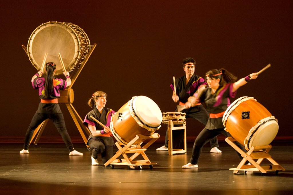 Group of performers energetically playing traditional Japanese taiko drums, captured mid-performance with vibrant stage lighting and expressive movements.