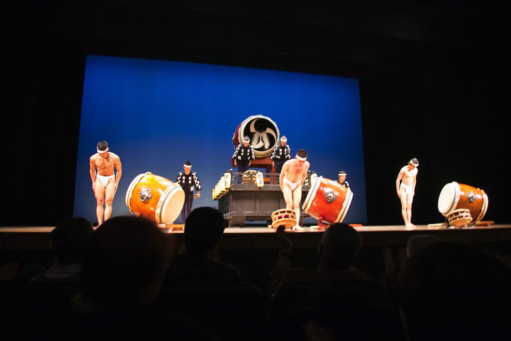 Taiko drummers performing on stage with large drums and traditional attire against a blue backdrop.
