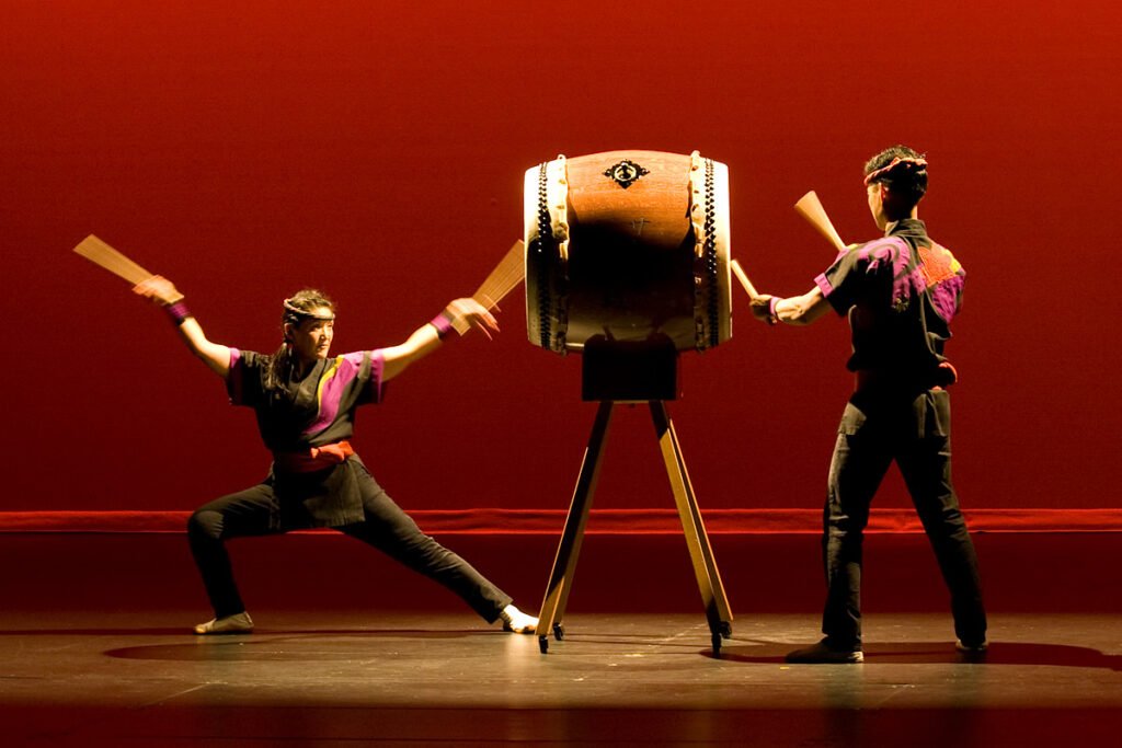 Two performers playing taiko drums with synchronized movements under dramatic red lighting