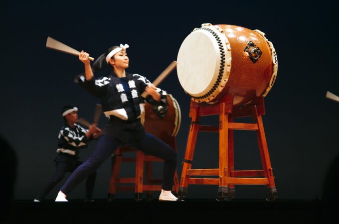 Enthusiastic performers playing large taiko drums on stage under bright lights