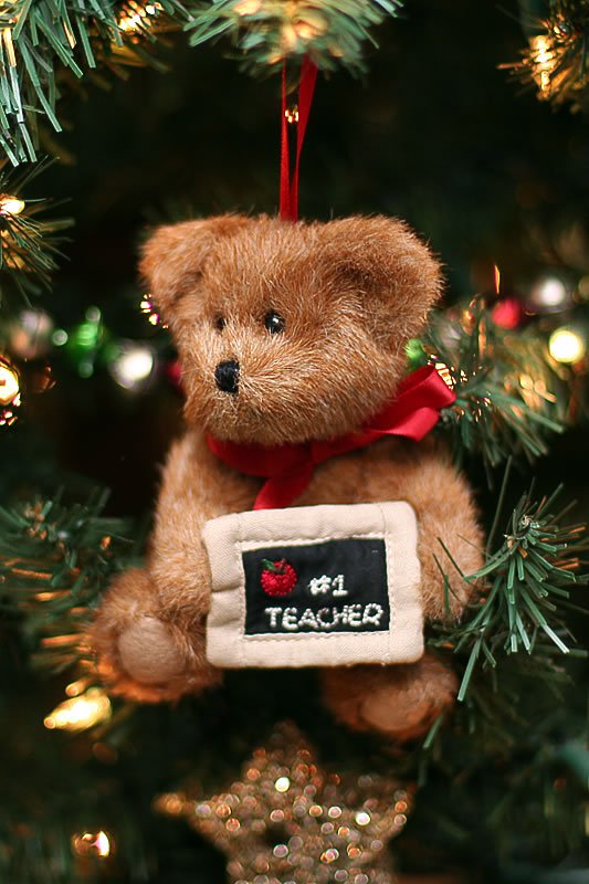 A charming teddy bear ornament with a red bow is hung on a Christmas tree. The bear holds a sign that reads '#1 Teacher'.