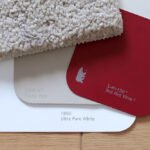A close-up view of a textured white carpet alongside red wine and ultra pure white color swatches on a wooden surface.