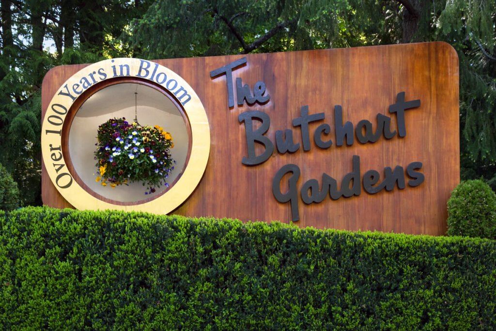A wooden sign for The Butchart Gardens featuring a hanging flower basket. The text 'Over 100 Years in Bloom' circles the floral display on the sign.