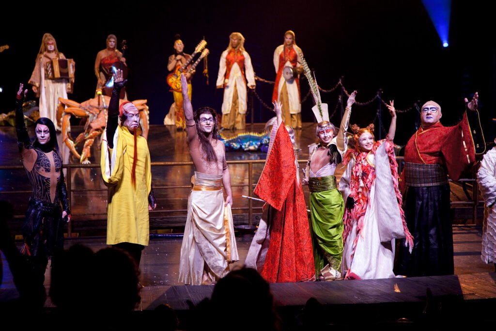 A group of theatrical performers in colorful outfits on a dimly lit stage with spotlights.