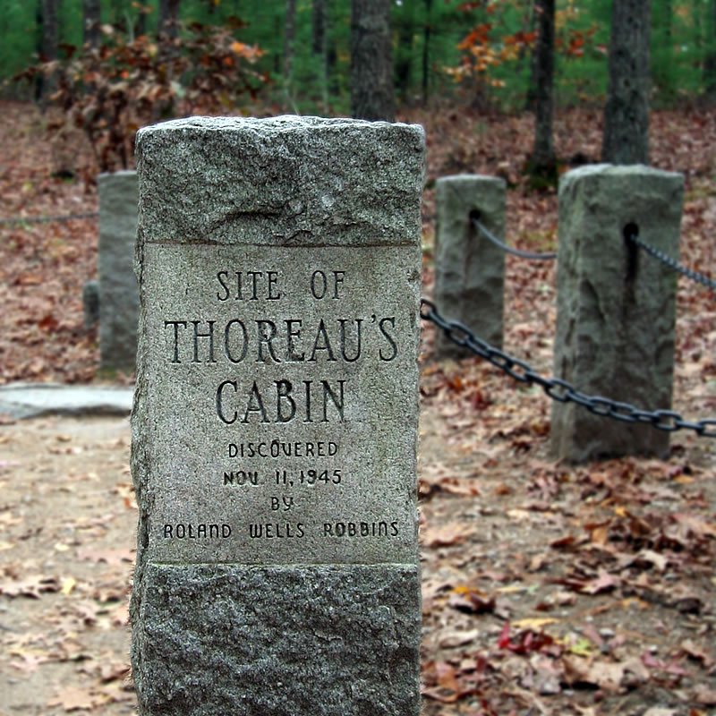 A stone marker in a forest clearing identifies the site of Thoreau's Cabin. The ground is covered in autumn leaves, surrounded by wooden posts connected by chains.
