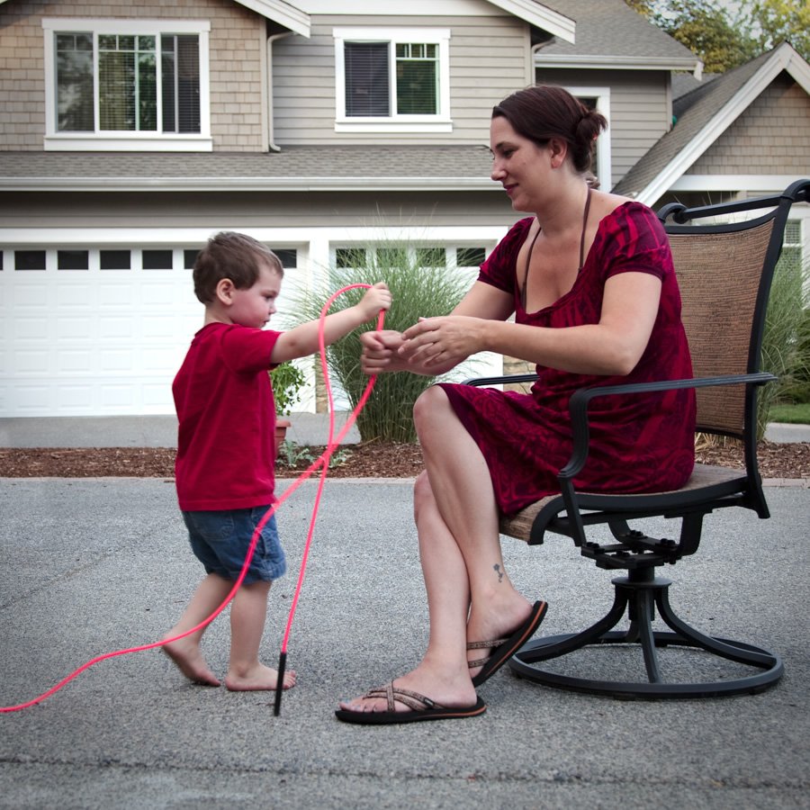 A woman in a red dress sits on a chair while a toddler in a red shirt plays with a pink hula hoop in a driveway.