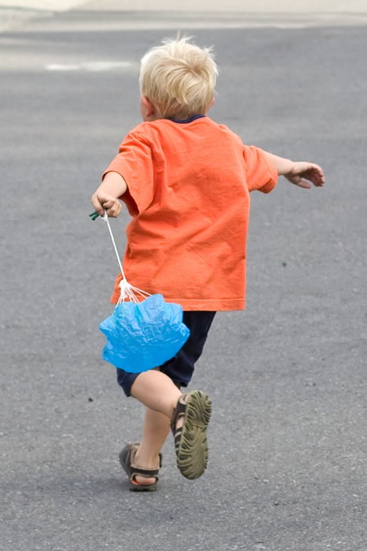 A young child with blonde hair in an orange shirt runs after a blue balloon on a paved surface.