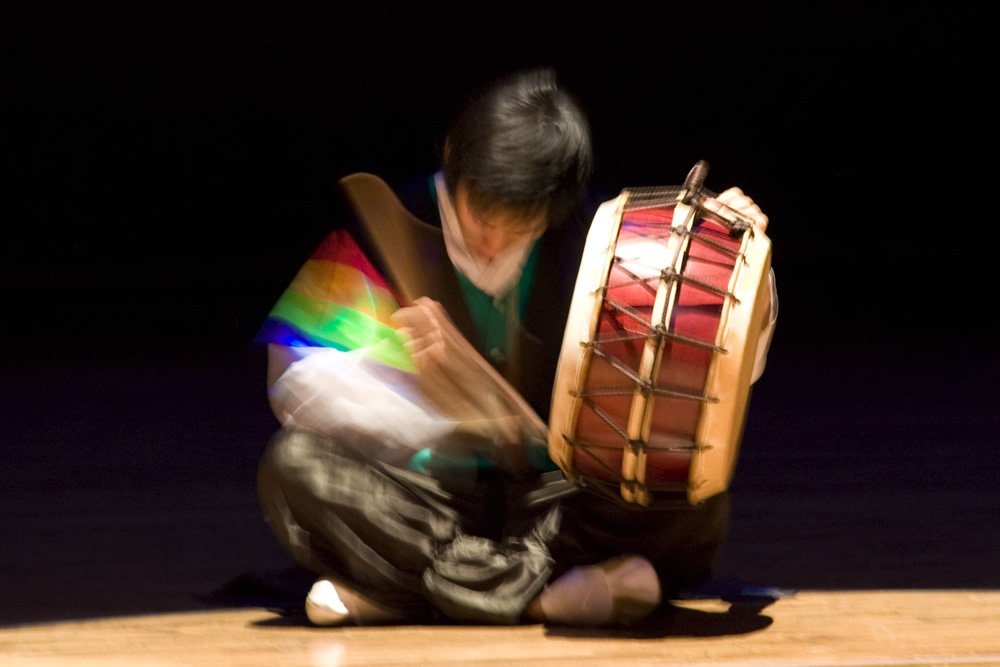 A performer on stage playing a traditional drum, illuminated by colorful stage lights.