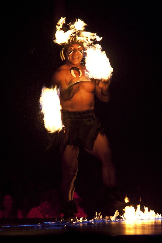 A performer is showcasing a traditional dance with fire during a nighttime event. Bright flames light up the area around the dancer.