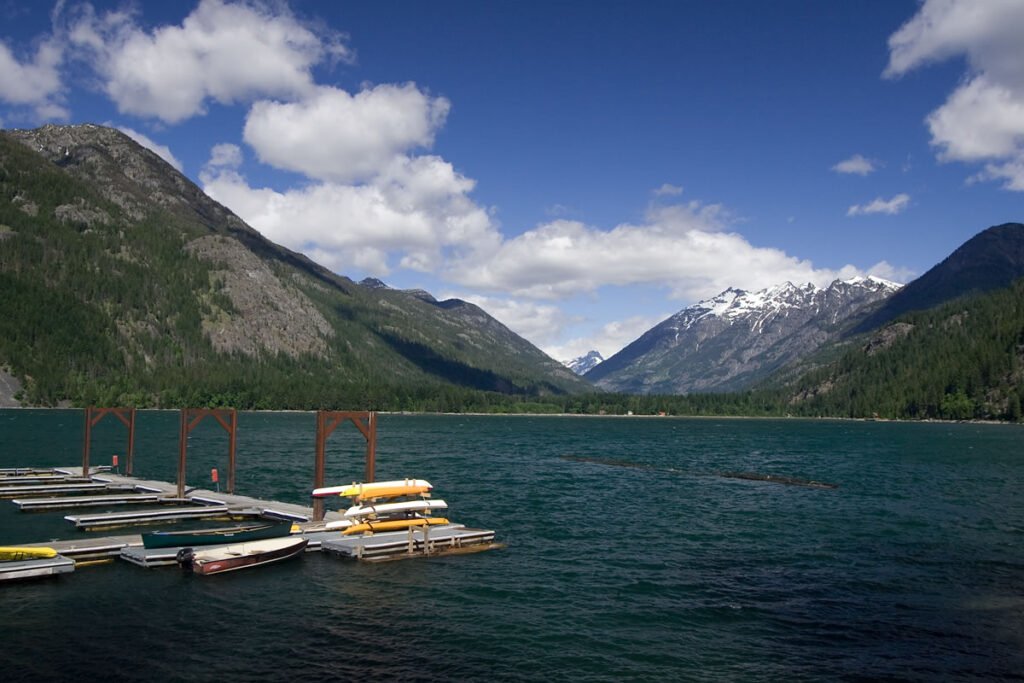 A serene lake surrounded by majestic mountains under a partly cloudy sky. Several kayaks are docked at a floating pier on the calm water.