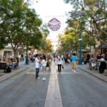 A vibrant street scene with people walking and sitting on benches under lush trees.