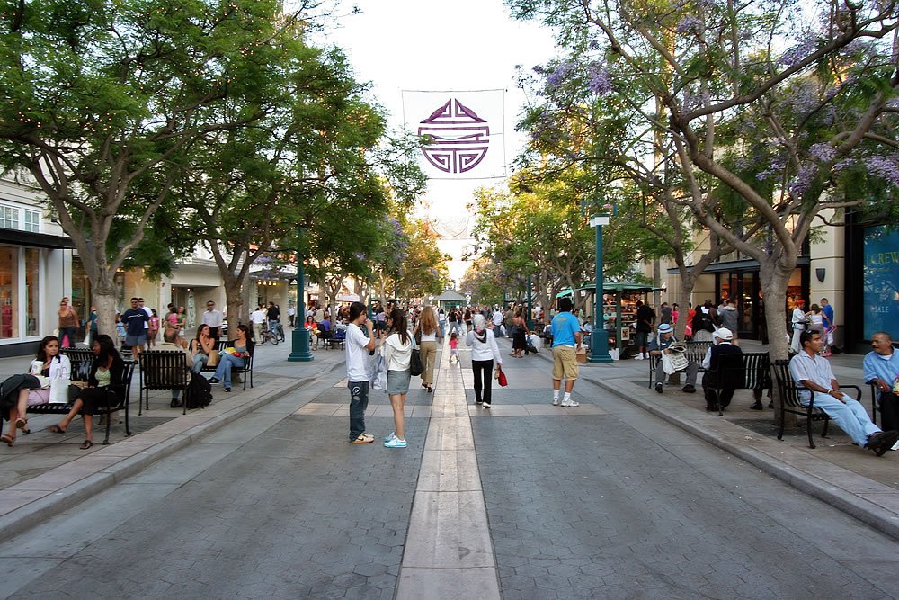 A vibrant street scene with people walking and sitting on benches under lush trees.