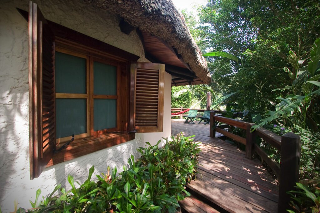 A cozy tropical shack with wooden window shutters and a thatched roof, surrounded by lush greenery along a wooden pathway.