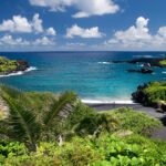 Iao Needle A breathtaking view of a tropical paradise with lush greenery, palm trees, and a pristine black sand beach. The turquoise waters of the Pacific Ocean stretch out under a bright blue sky dotted with fluffy white clouds.