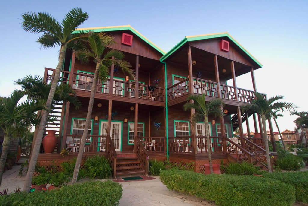 A stunning two-story beachfront villa surrounded by lush palm trees and greenery, featuring vibrant red and green accents on its wooden exterior.