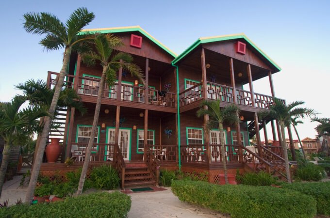 A stunning two-story beachfront villa surrounded by lush palm trees and greenery, featuring vibrant red and green accents on its wooden exterior.