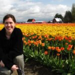 U2 A woman smiles in front of colorful tulip fields. The scene includes vibrant yellow and orange tulips with farm buildings in the background.