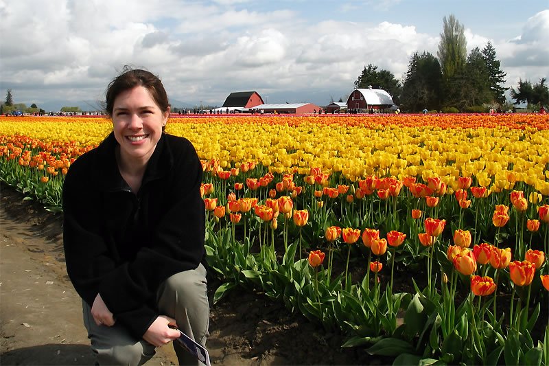 A woman smiles in front of colorful tulip fields. The scene includes vibrant yellow and orange tulips with farm buildings in the background.