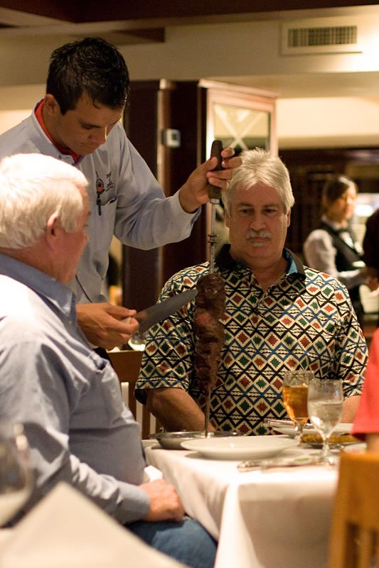 A close-up of a waiter preparing food at a table with two diners, one wearing a patterned shirt, in a warmly lit restaurant setting.