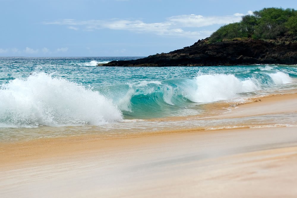 Vibrant turquoise waves breaking on a sandy beach with a rocky, green-covered cliff in the background under a clear blue sky.