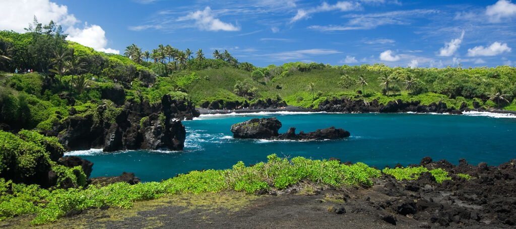 A breathtaking view of turquoise waters surrounded by lush green hills and rocky formations under a bright blue sky with fluffy white clouds.